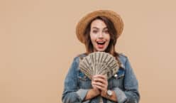 Portrait of a smiling young girl in summer clothes showing money banknotes and looking at camera isolated over beige background for the concept "What is passive income?"